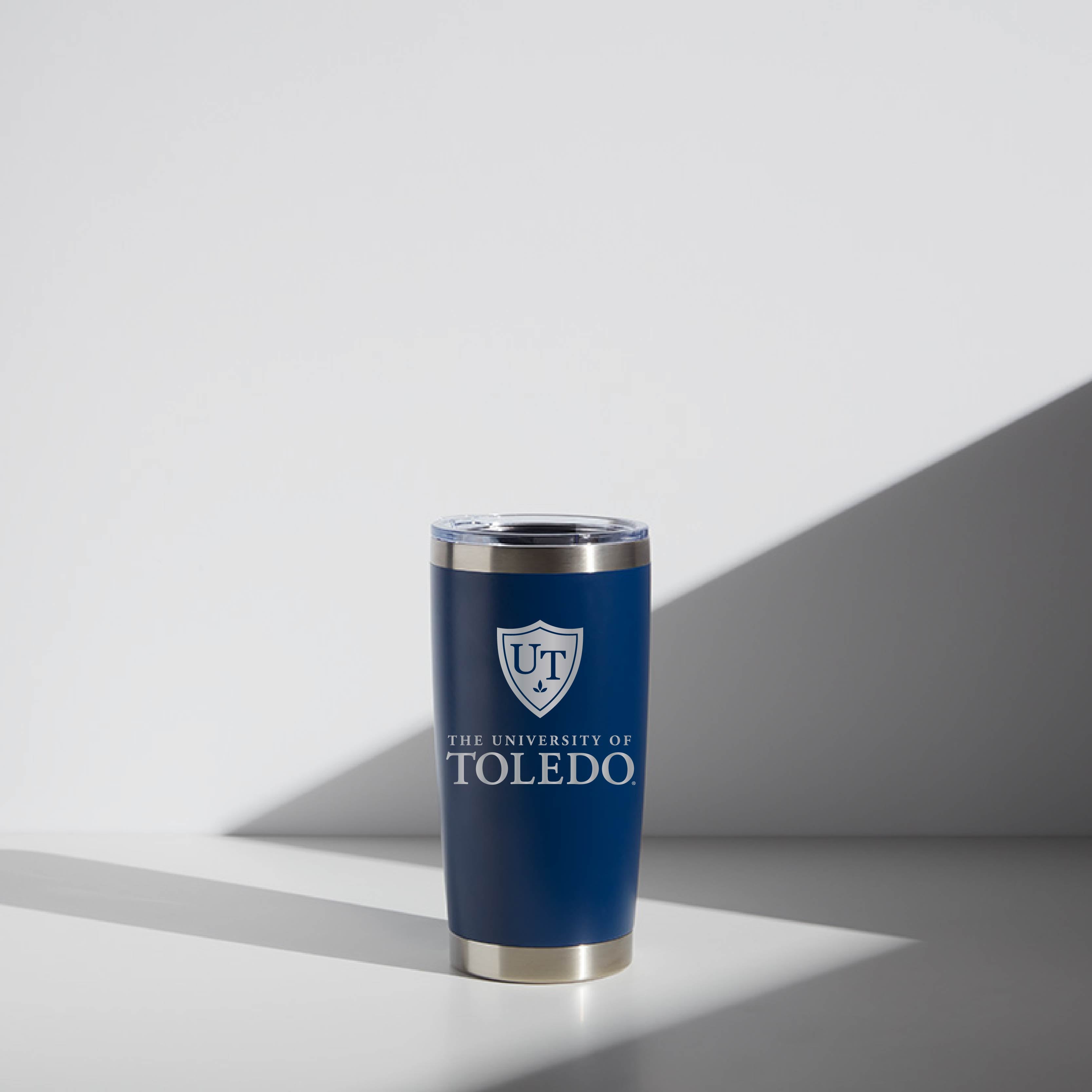 Alt text: "University of Toledo branded 20 oz WATR Tumbler in navy blue with a silver lid, featuring the UT shield logo and name on the front. It stands on a light gray surface with a soft shadow cast to the