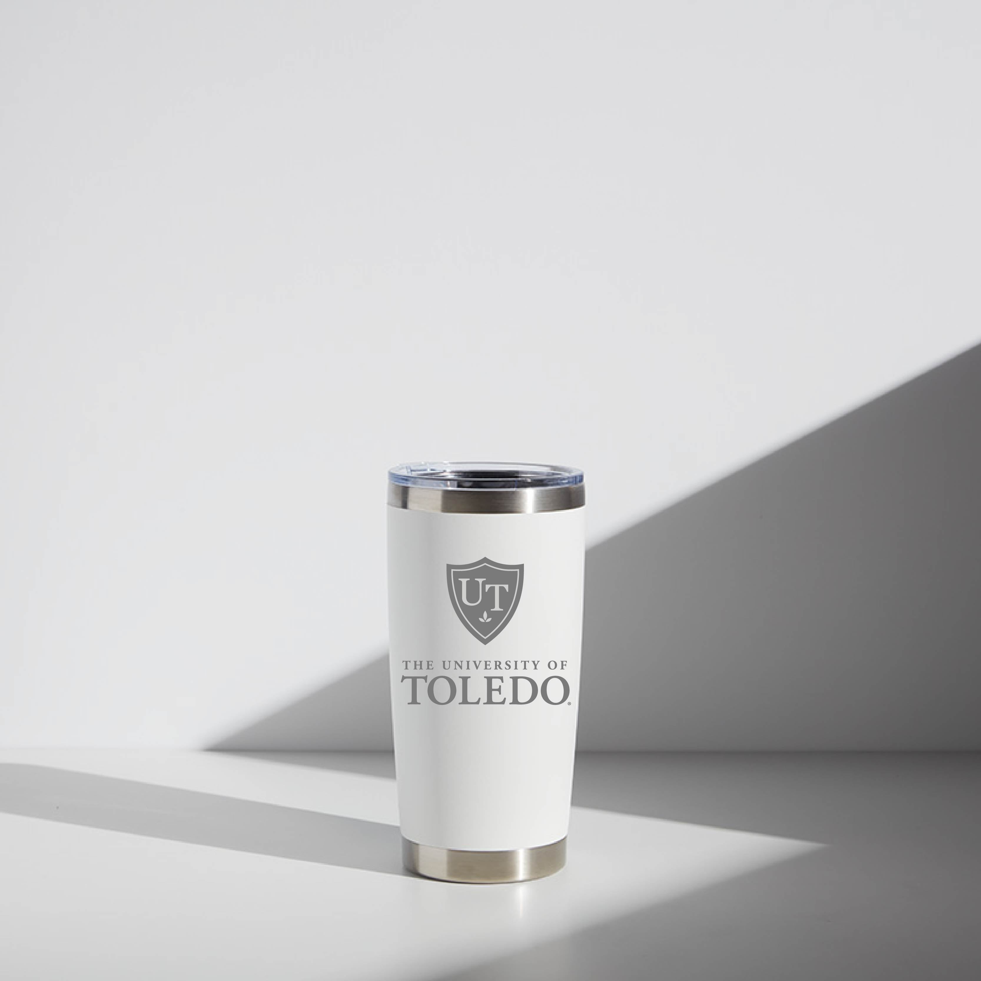 A 20 oz WATR tumbler in white and silver colors featuring the University of Toledo logo in a shield design on its smooth surface, photographed in natural light with a soft shadow to its right.