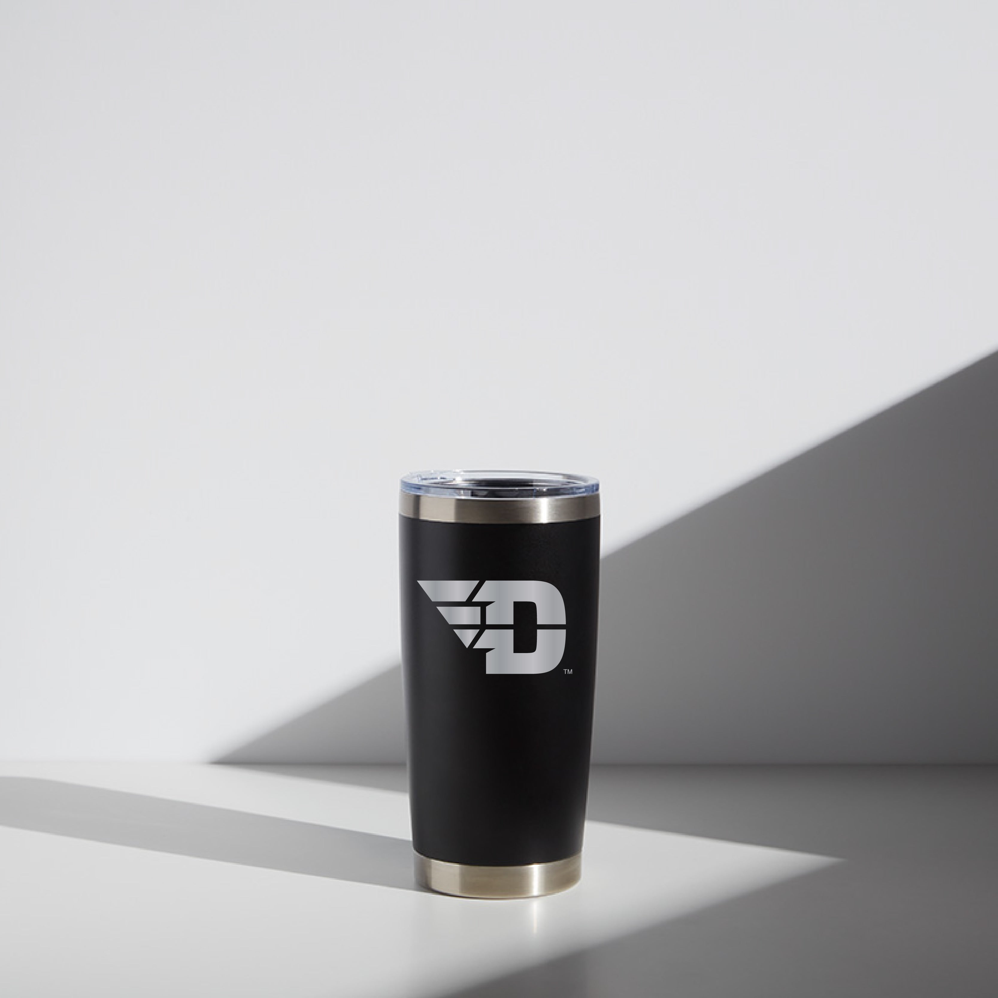 Alt text: "20 oz black WATR tumbler with the Dayton 'Flying D' logo on the front, featuring a silver trim at the bottom and a clear lid, set against a minimalist gray and white background."