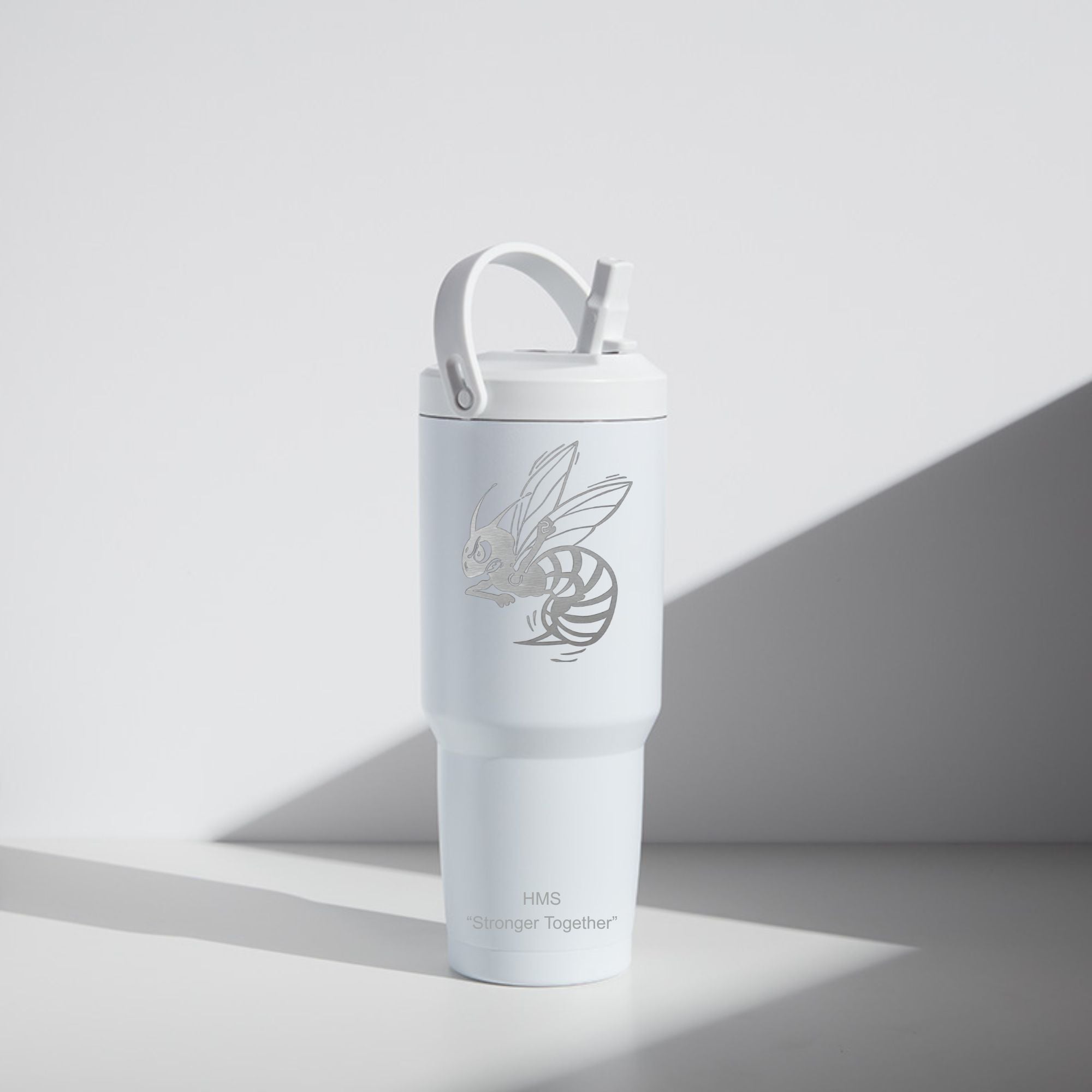 A white insulated travel tumbler with a matching lid and straw, featuring a gray bee graphic and the text 'HMS Stronger Together' near the bottom, photographed on a light gray surface with a strong diagonal shadow.