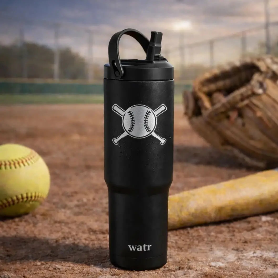 Black insulated stainless steel water bottle with engraved baseball and crossed bats design, sitting on a dirt baseball field with a softball, glove, and bat in the background