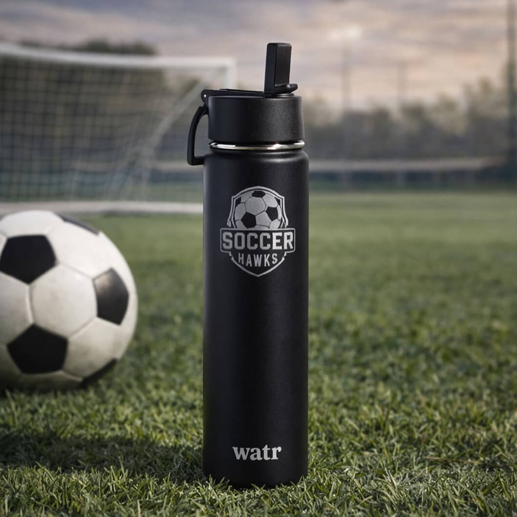 Black insulated stainless steel water bottle with engraved Soccer Hawks logo, standing upright on a grass soccer field with a soccer ball and goal net in the background