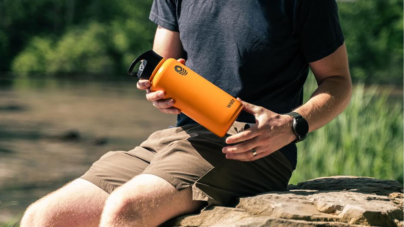 Man sitting on a rock near water, holding an orange We Are The Ripple (WATR) BPA-free water bottle.