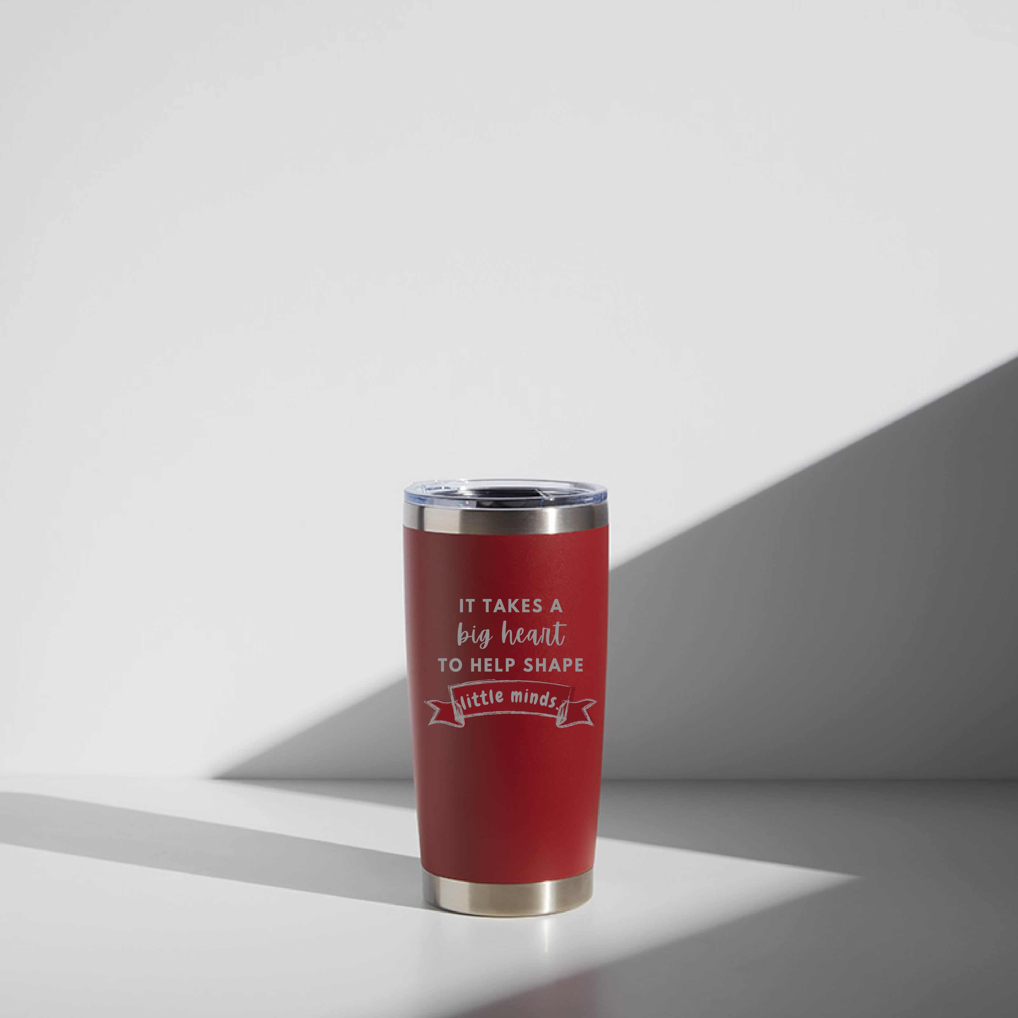 Red 20 oz stainless steel tumbler with a personalized message, "It takes a big heart to help shape little minds," ideal for teacher gifts, displayed in a softly lit setting with a geometric shadow.