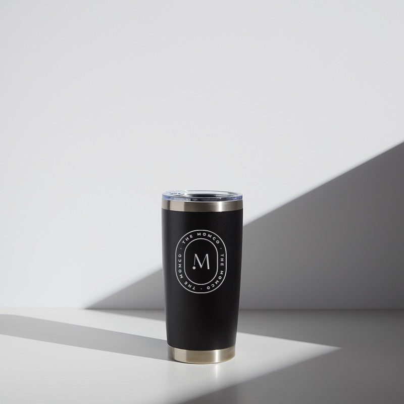 Alt text: "20 oz MomCo WATR stainless steel tumbler in black with a clear lid, featuring a white logo with the letter 'M' in a circle on the front, set against a light background with a soft shadow."