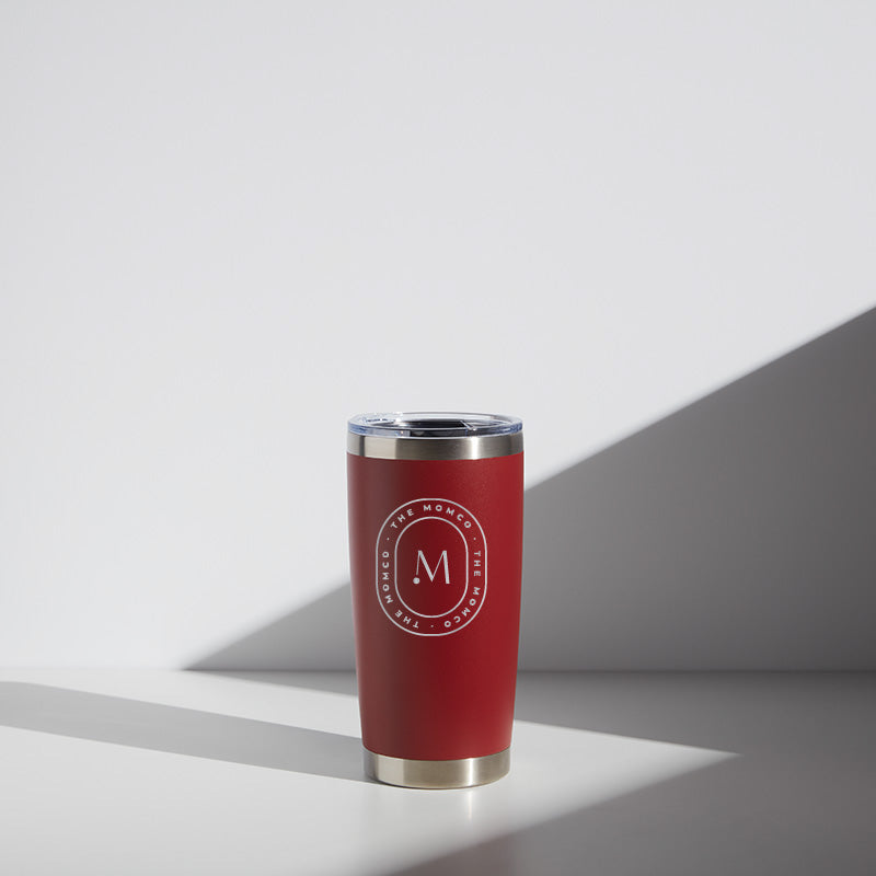 Alt text: "Red 20 oz MomCo WATR stainless steel tumbler with clear lid, featuring a white circular logo with the letter 'M' centered, standing on a white surface with a shadow to the right."