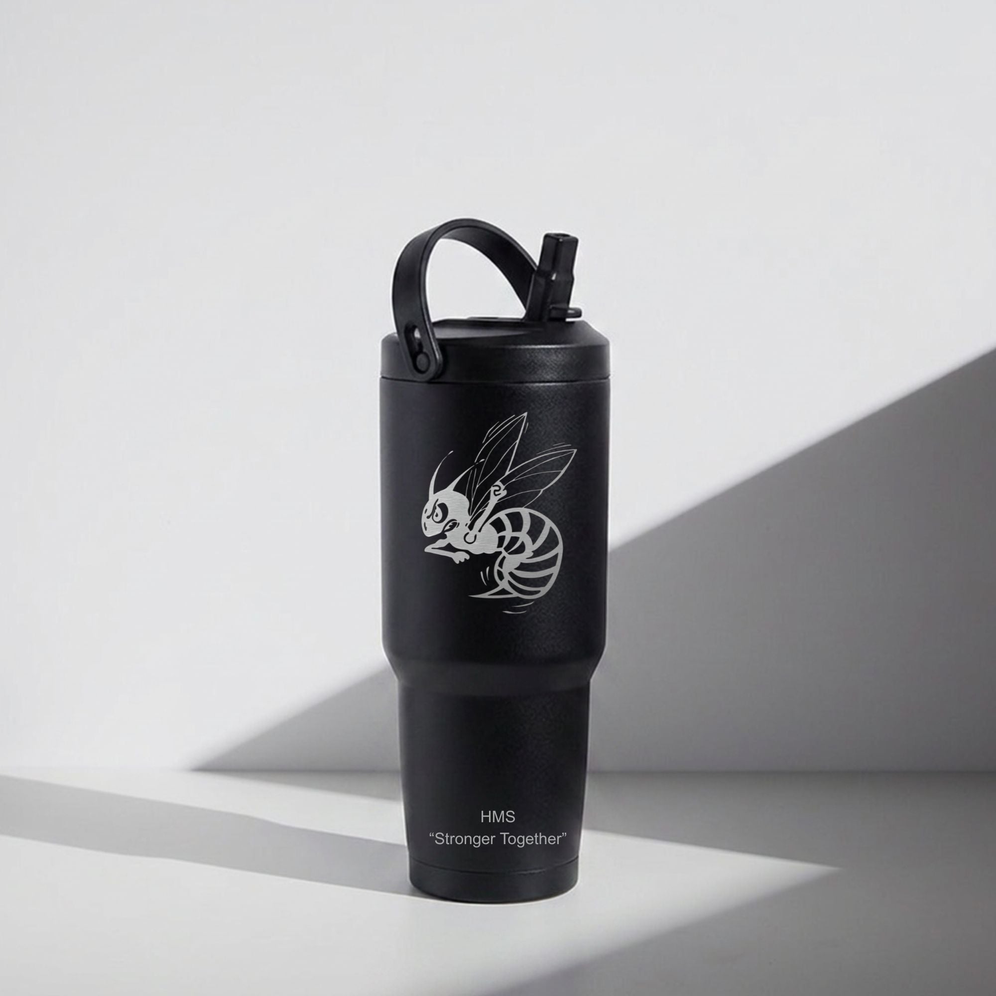A matte black insulated travel tumbler with a carry loop lid and straw, featuring a white graphic of a bee and the text 'HMS Stronger Together' on a clean white background with dramatic shadows.