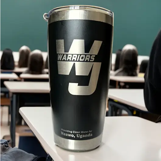 Custom engraved black Warriors stainless steel water bottle on a classroom desk, inscribed with "Providing Clean Water for Nsawo, Uganda"
