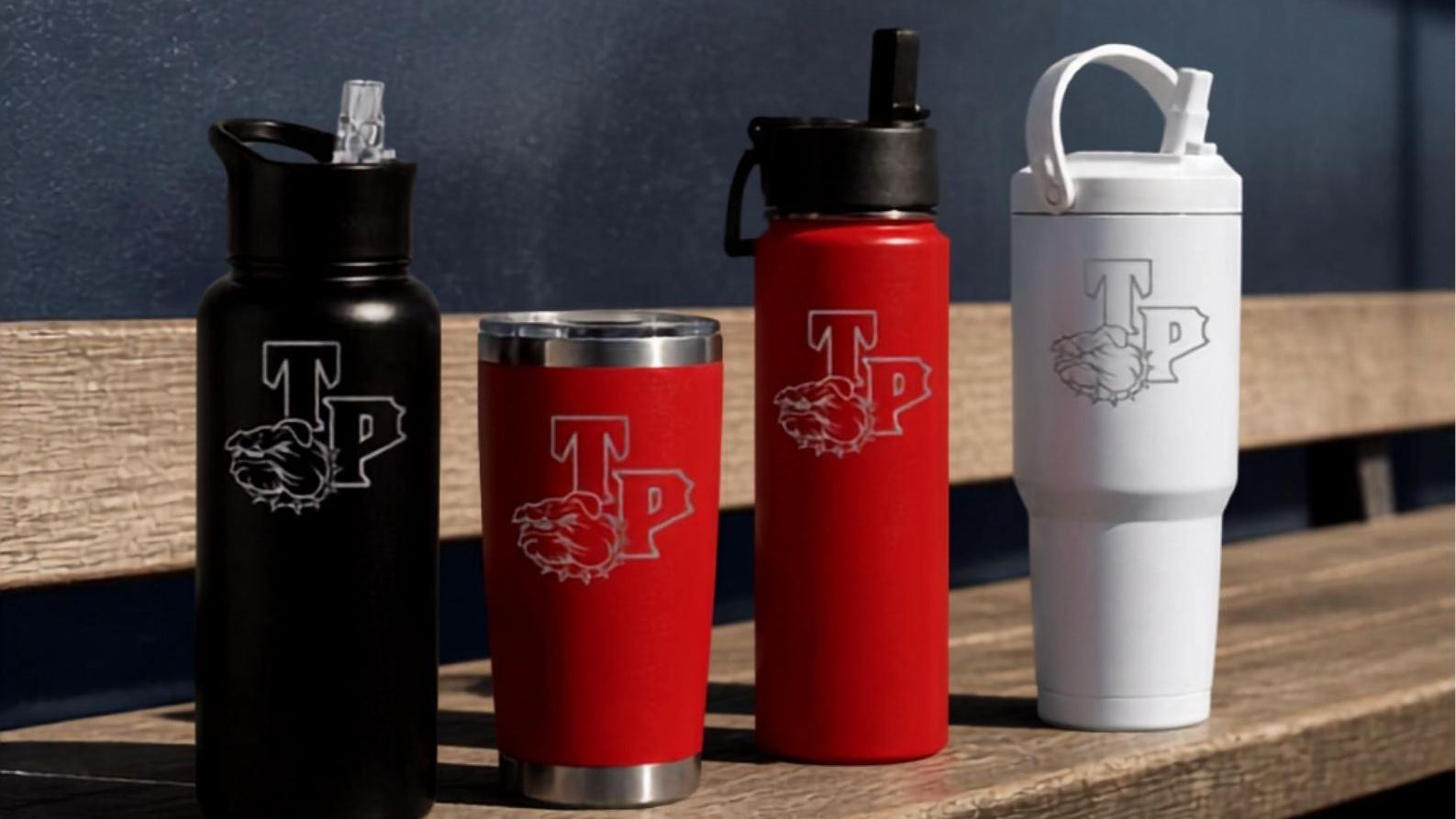 A promotional graphic showing three branded insulated tumblers and a water bottle lined up on a wooden bench: a black bottle, a red tumbler, and a red water bottle, plus a white tumbler. Large text reads 'BUILT FOR THE GAME' with smaller copy 'HYDRATE.