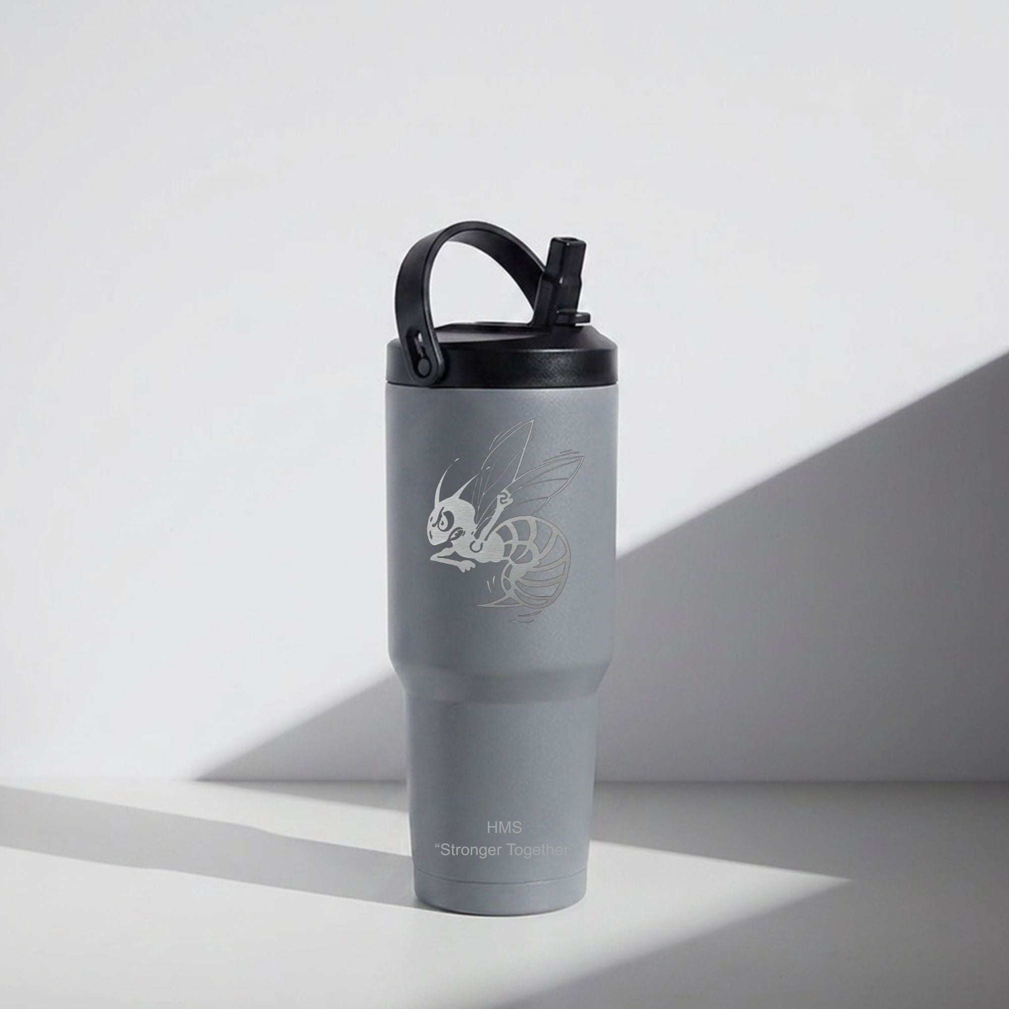 A matte gray insulated tumbler with a black lid and carry loop, featuring a white bee graphic and the text 'HM8 Stronger Than' near the bottom, photographed on a white surface with a diagonal shadow.