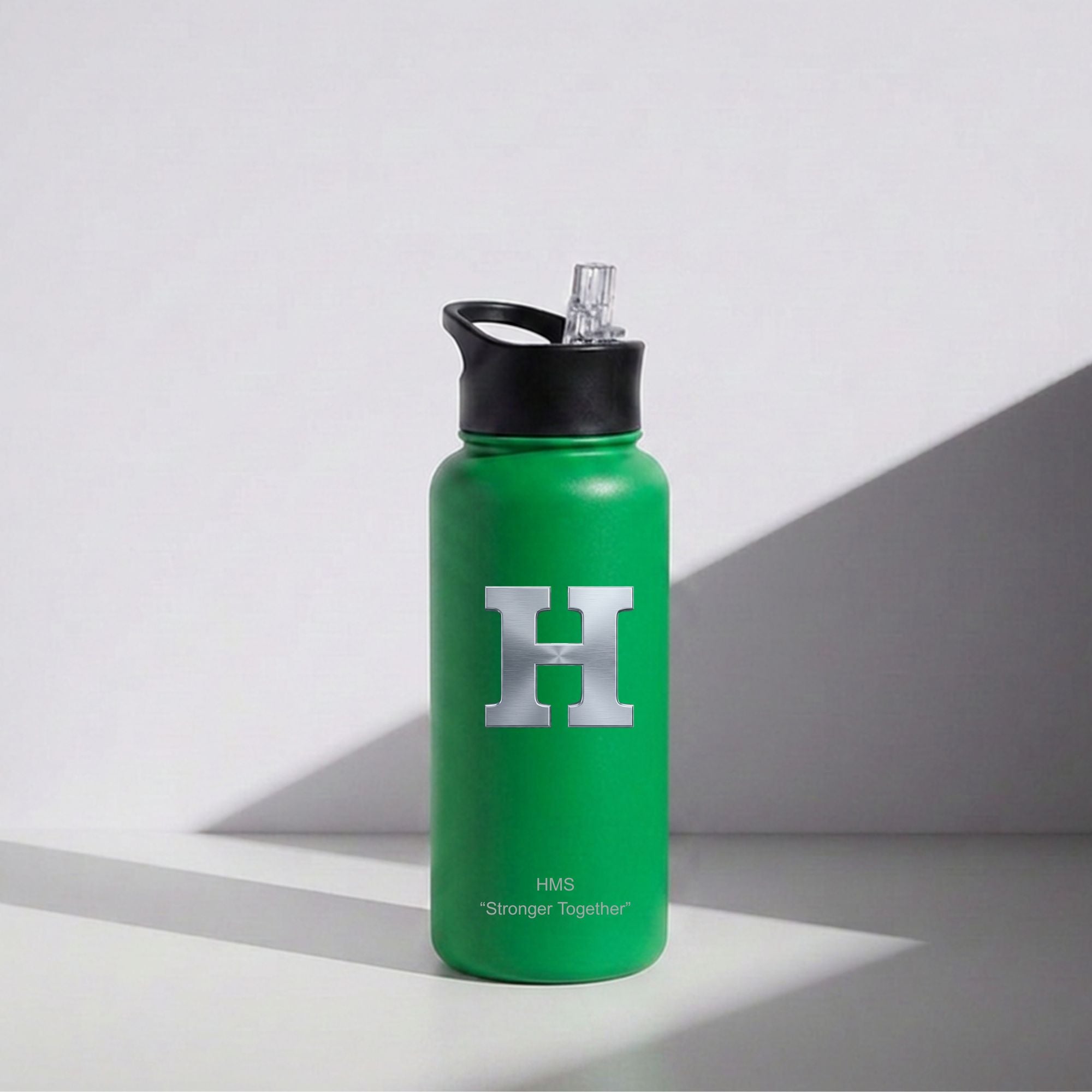 A green insulated water bottle with a black lid and clear straw, featuring a large silver 'H' logo and small 'FMS Stronger Together' text, photographed on a light gray surface with a strong diagonal shadow.
