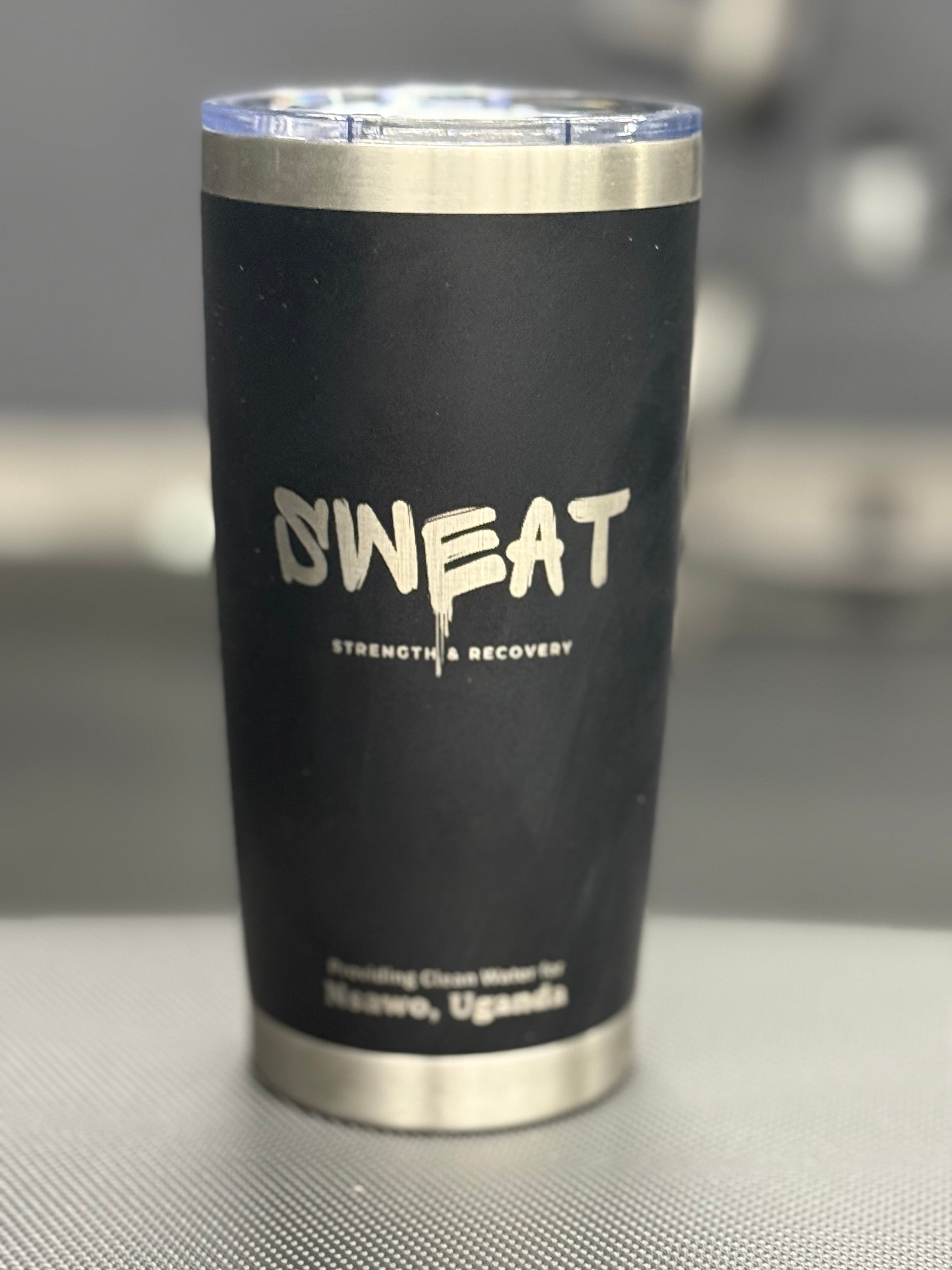 Alt text: "20 oz custom black stainless steel tumbler with a silver rim and a clear lid. The tumbler is branded with the white word 'SWEAT' and the slogan 'Strength Recovery' underneath. Additional text includes '