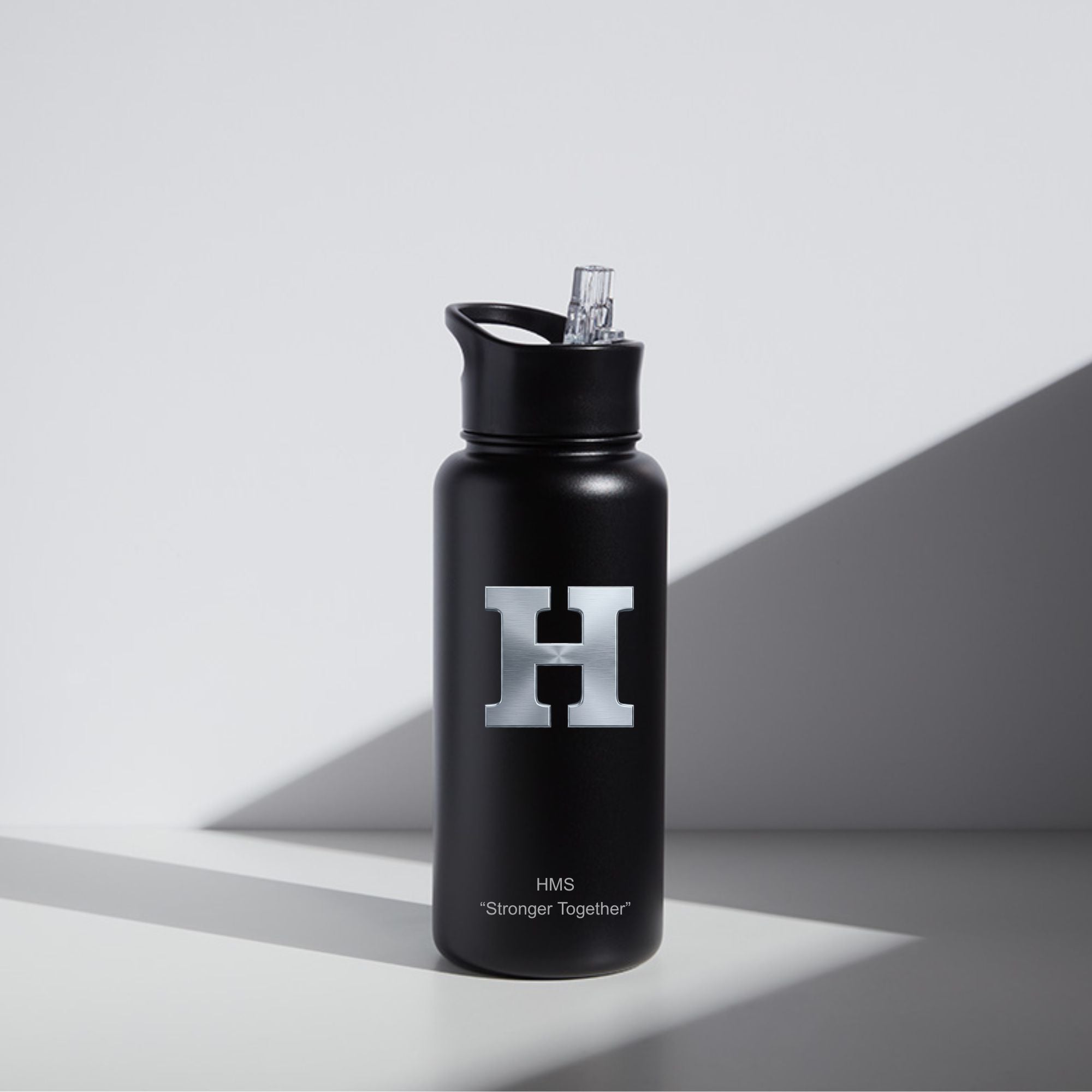 A matte black water bottle with a silver 'H' logo and 'HMS Stronger Together' text, standing on a light gray surface with dramatic diagonal shadows.